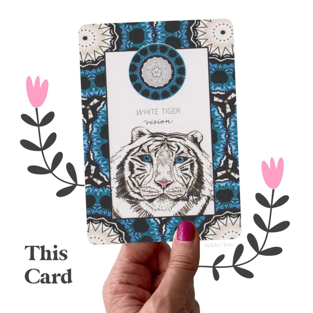 A female hand holding the White Tiger Oracle Cards from Animal Kin Deck by Sarah Wilder. The image also contains two pink & black illustrated flowers and the words 'This Card" and bakkehus logo.