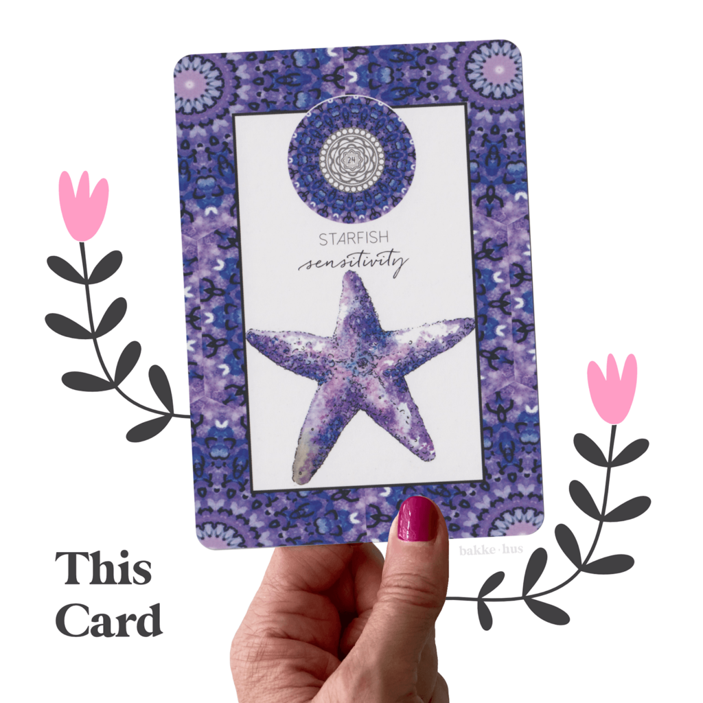 A female hand holding the Starfish Oracle Card from Animal Kin Deck by Sarah Wilder. The image also contains two pink & black illustrated flowers and the words 'This Card" and bakkehus logo.