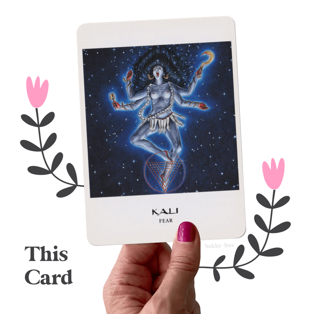 A female hand holding the Kali Oracle Cards from The Goddess Oracle deck by Amy Sophia Marashinsky. The image also contains two pink & black illustrated flowers and the words 'This Card" and bakkehus logo.