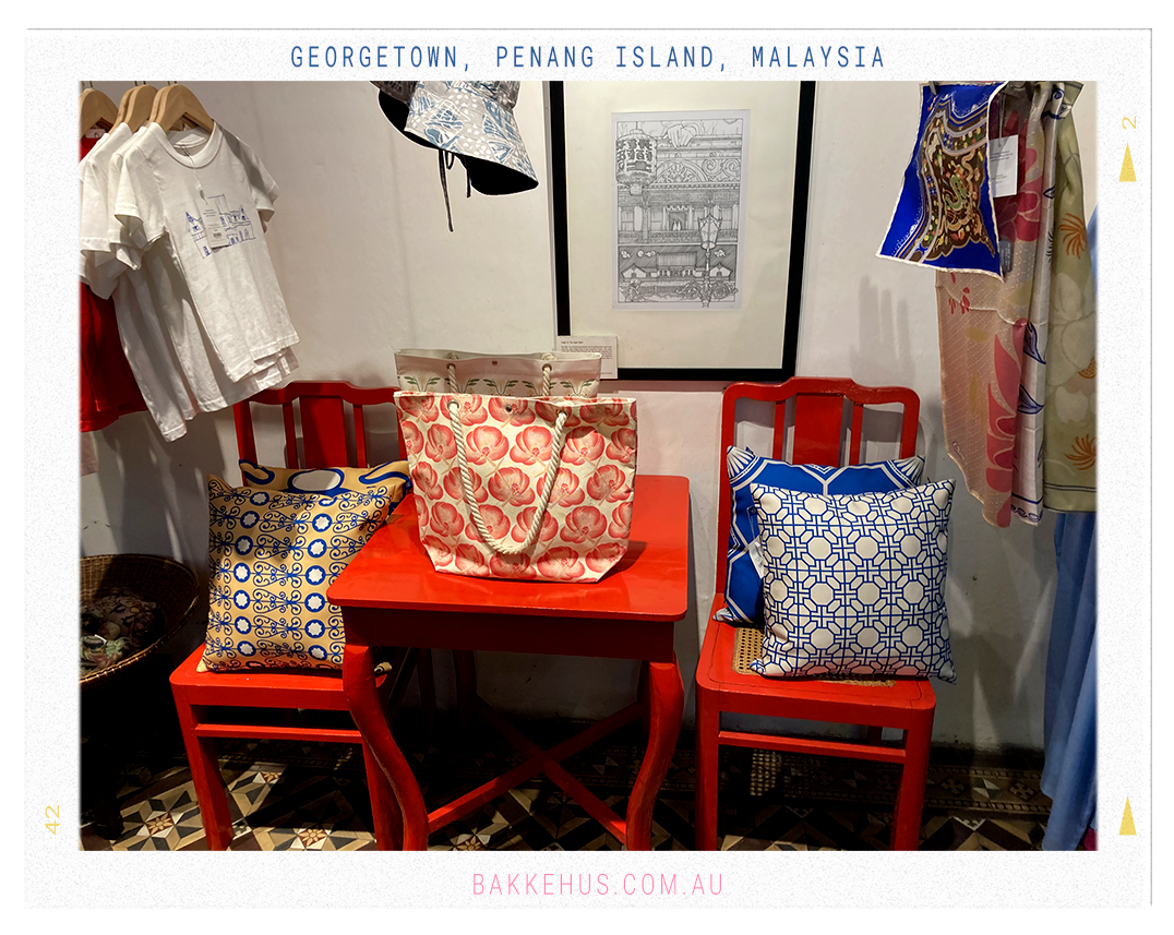 Colorful textile and souvenir display inside the Blue Mansion gift shop in George Town, Penang, Malaysia, featuring patterned pillows, printed tote bags, architectural-themed T-shirts, and silk scarves arranged against bold red furniture. © Heidi Isaksen-Loxton for bakkehus.com