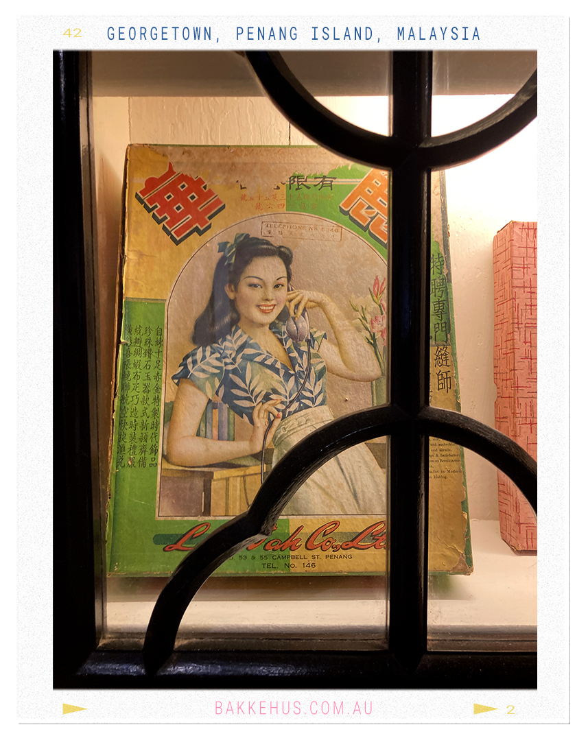 Framed view of a vintage Chinese advertisement poster featuring a smiling woman in a tropical blouse, displayed behind decorative cupboard door at the Blue Mansion in George Town, Penang, Malaysia—offering a glimpse into mid-20th-century local design and culture. © Heidi Isaksen-Loxton for bakkehus.com