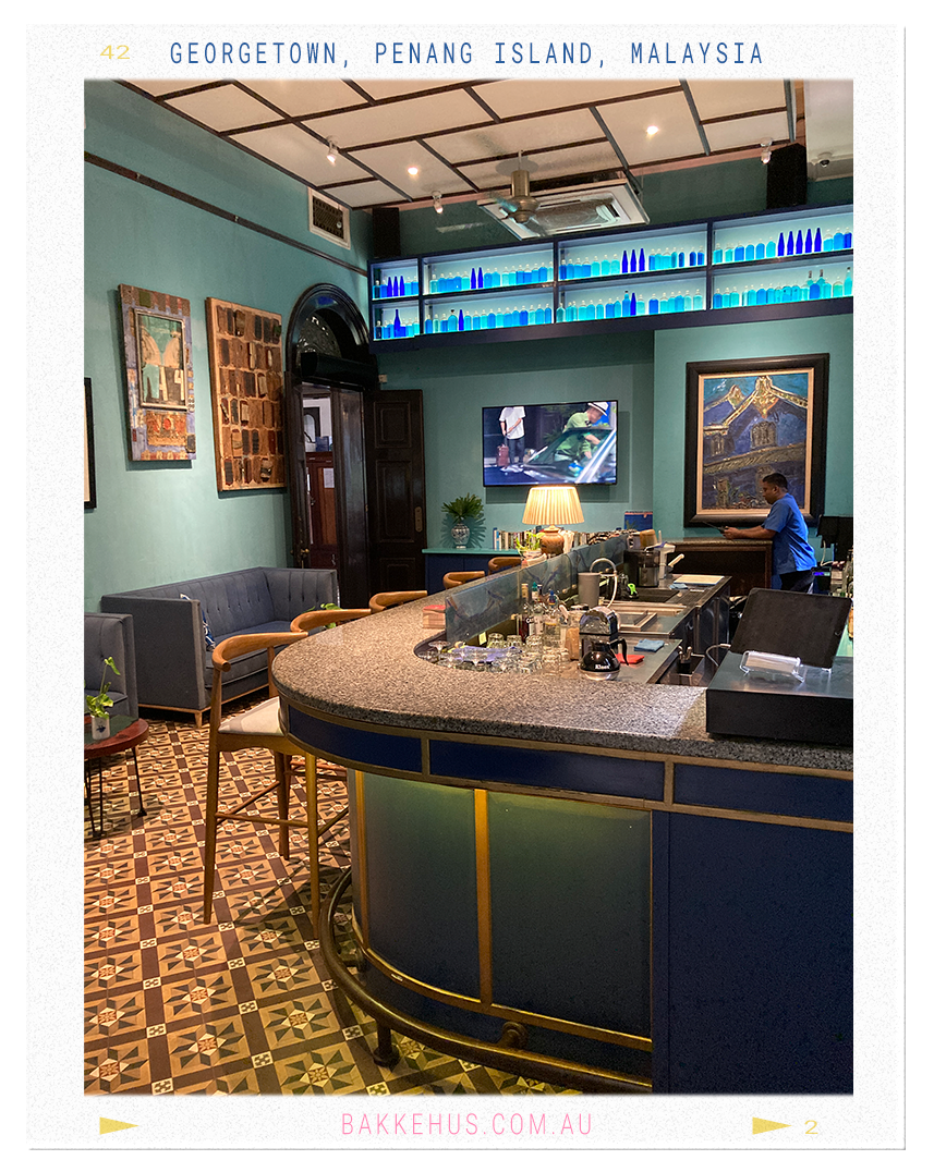 Interior of the Blue Mansion bar in George Town, Penang, Malaysia, featuring a curved granite-topped counter, heritage floor tiles, teal walls, and blue glass bottle displays—blending colonial elegance with Peranakan charm. © Heidi Isaksen-Loxton for bakkehus.com