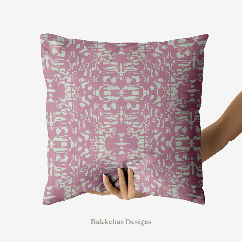 A hand holds up a square cushion printed with Abstract Inked Lace, a large-scale mirrored lace pattern in pale sage green on dusty mauve.