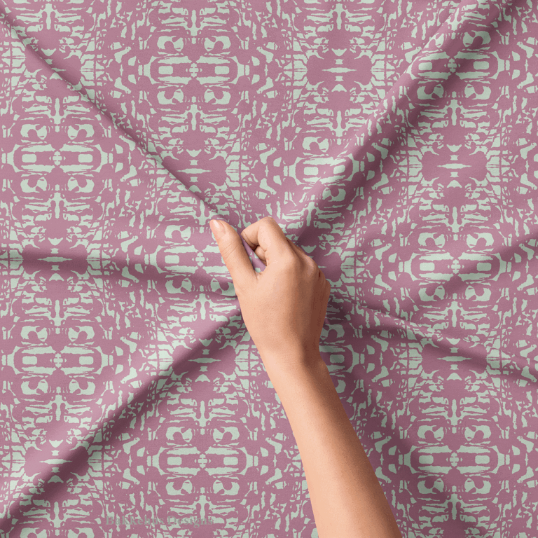 Close-up of a hand lifting Abstract Inked Lace fabric, showing the rough, ink-blot edges of the pale sage green shapes on dusty mauve.