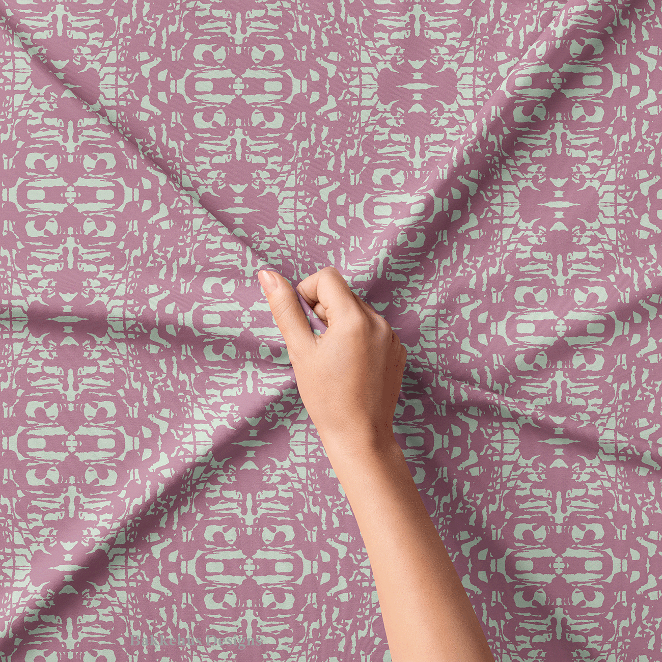 Close-up of a hand lifting Abstract Inked Lace fabric, showing the rough, ink-blot edges of the pale sage green shapes on dusty mauve.