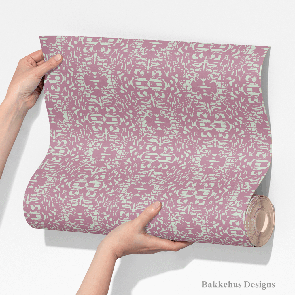 Two hands unroll a length of Abstract Inked Lace wallpaper, showing the large mirrored lace shapes in pale sage green on a dusty mauve background.