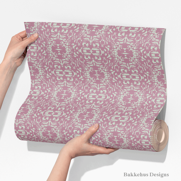Two hands unroll a length of Abstract Inked Lace wallpaper, showing the large mirrored lace shapes in pale sage green on a dusty mauve background.
