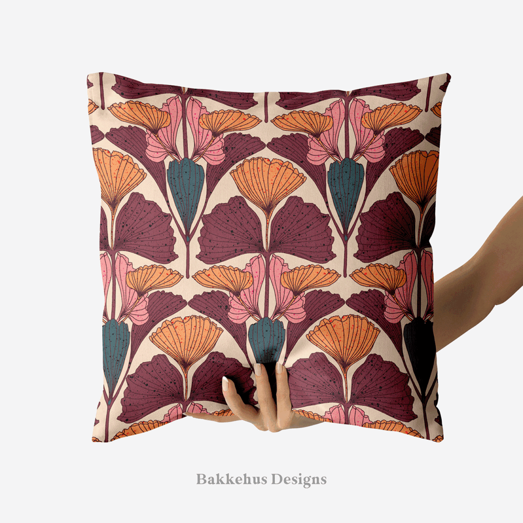 A hand holds up a square cushion printed with Art Nouveau Ginkgo Fan, a pattern of mauve, burnt orange and dusty pink ginkgo leaves with deep teal buds on a warm beige background.