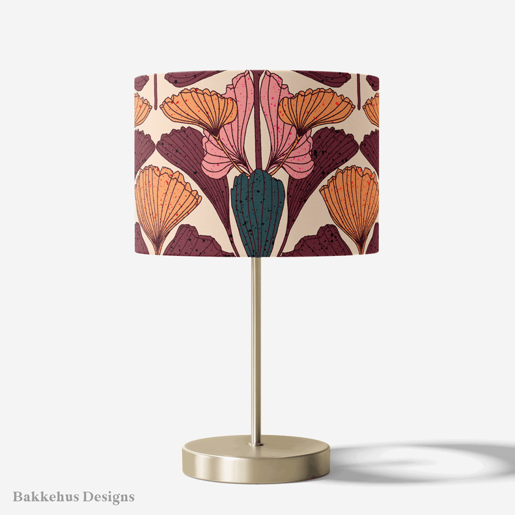 Table lamp with a brass stand and a round drum shade covered in the Art Nouveau Ginkgo Fan pattern in plum, burnt orange, pink and teal on beige.