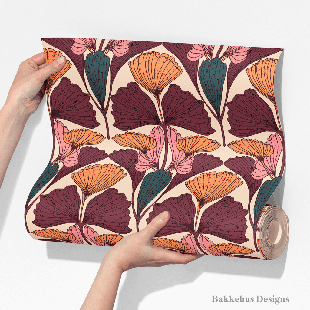 Two hands unroll a length of Art Nouveau Ginkgo Fan wallpaper, showing rows of plum, burnt orange and dusty pink ginkgo fans with teal buds on warm beige.