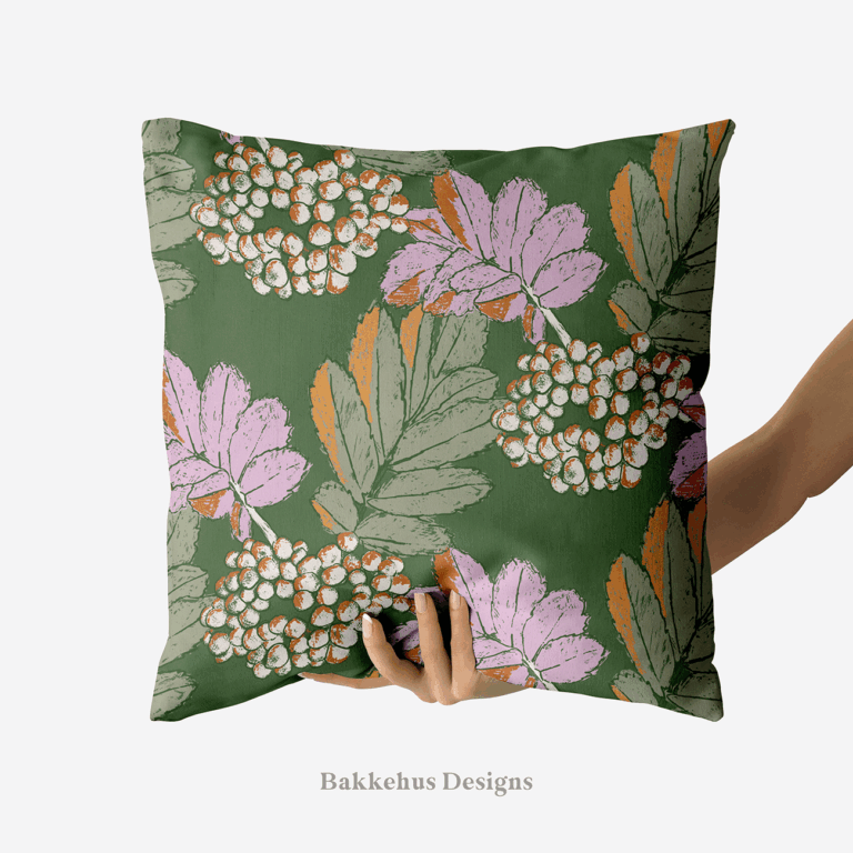 A hand holds up a square cushion printed with Bramble & Berry Botanical, a large-scale pattern of berry clusters and leafy branches in lilac and cream on forest green.