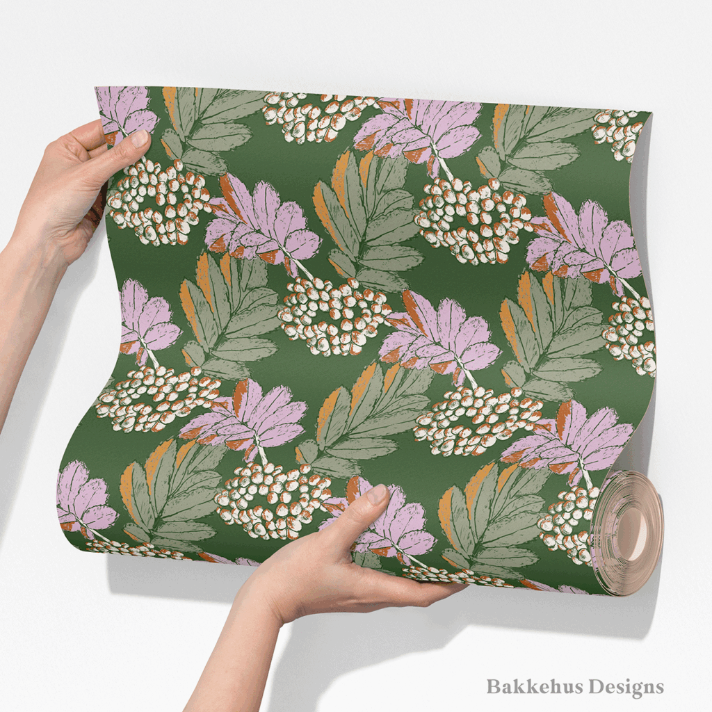 Two hands unroll a length of Bramble & Berry Botanical wallpaper, showing large berry clusters and leafy branches in lilac and cream on forest green.