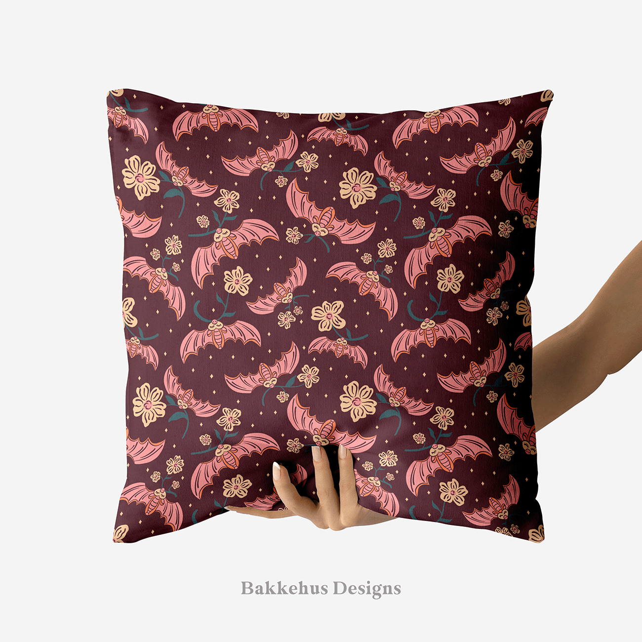 A hand holds up a square cushion printed with Cute Flower Bats: sleepy pink bats carrying sprigs of cream flowers across a dark plum night.