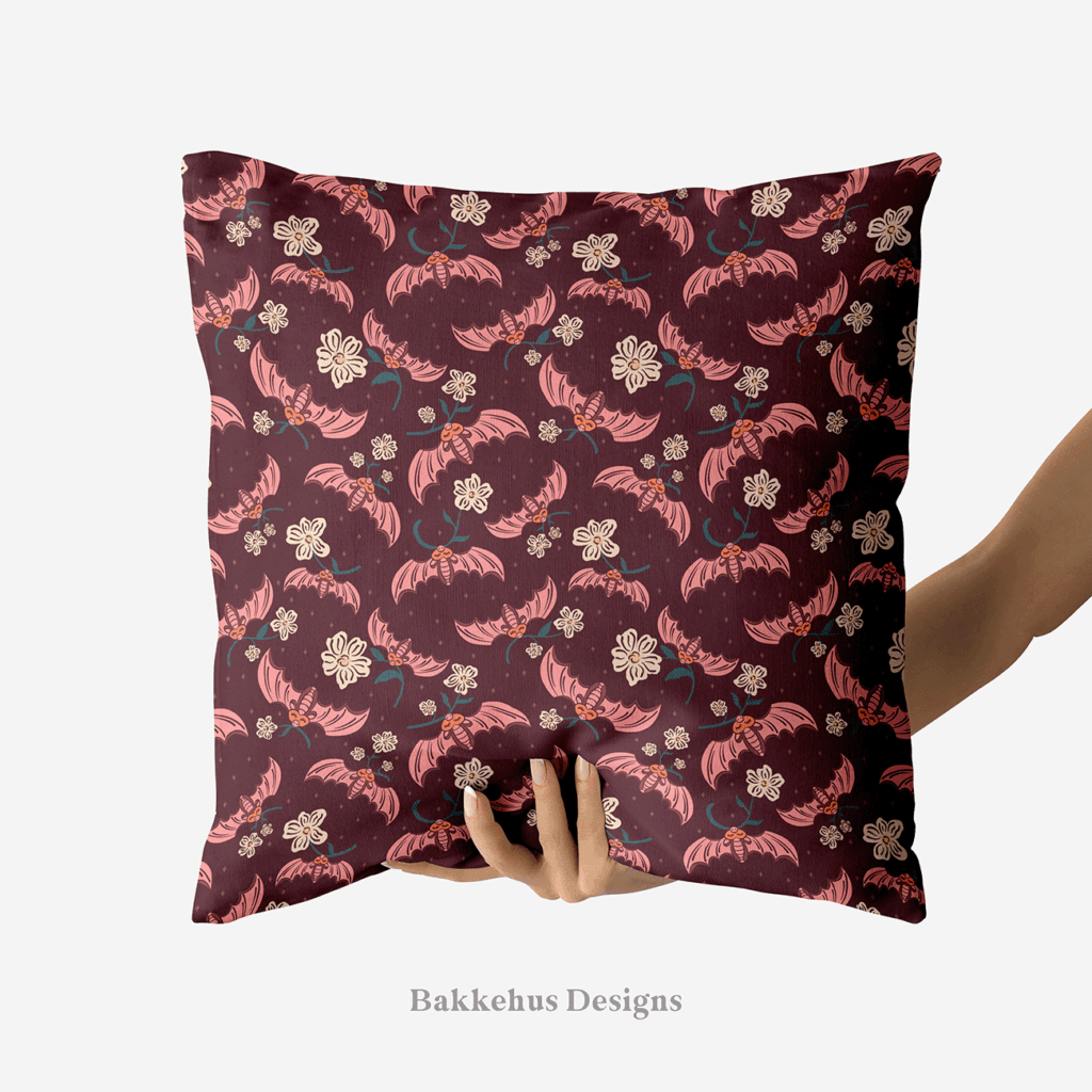 A hand holds up a square cushion printed with Cute Flower Bats: sleepy pink bats carrying sprigs of cream flowers across a dark plum night.