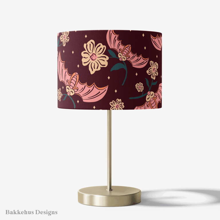 Table lamp with a brass stand and a round drum shade covered in Cute Flower Bats, with large pink bats and cream flowers on dark plum.