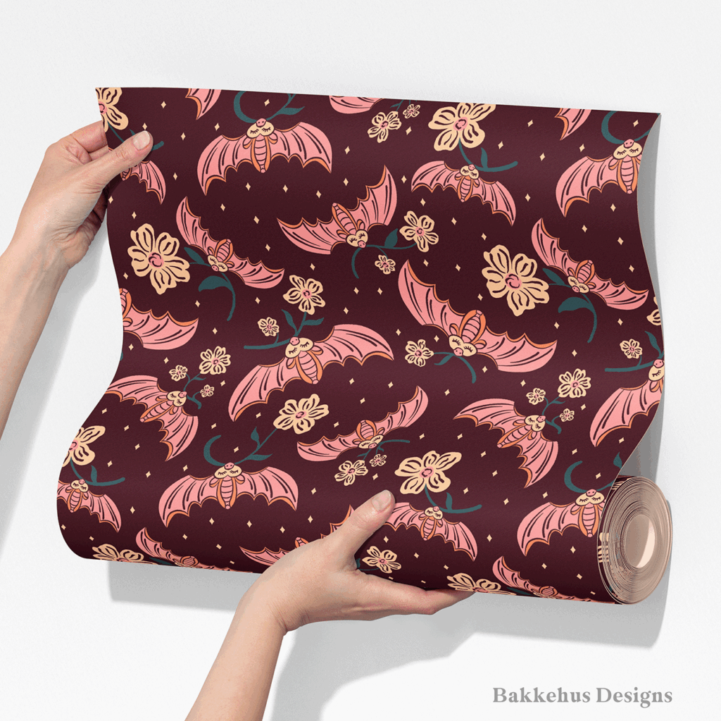 Two hands unroll a length of Cute Flower Bats wallpaper, showing pink bats and cream flower sprigs repeating across a dark plum ground.