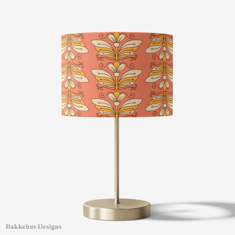 Table lamp with a brass stand and a round drum shade covered in the Retro Flower Moth Stripe pattern, with cream and gold flower moths on coral.