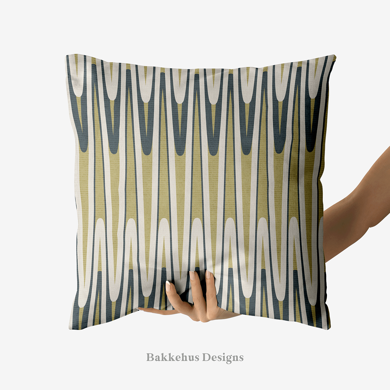 A hand holds up a square cushion printed with Retro Wavy Stripe, a vertical mid-century pattern of elongated looping waves in olive green and charcoal on a warm cream ground.